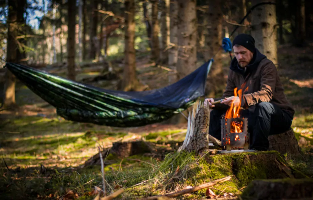a man sitting next to a fire with a warbonnet hammock with an attached green wookie underquilt hanging in the background